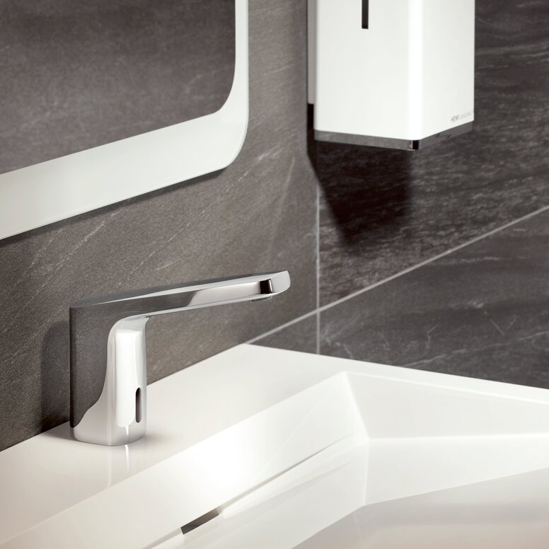 A chrome-plated touchless tap on a white washbasin against a backdrop of dark grey stone tiles