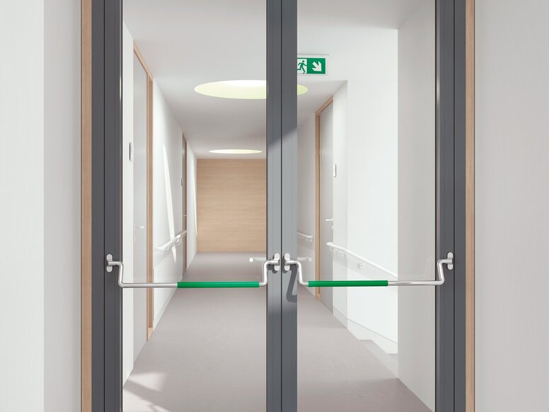 Double-leaf glass door with a grey frame and panic bars with green grip surfaces in a white corridor