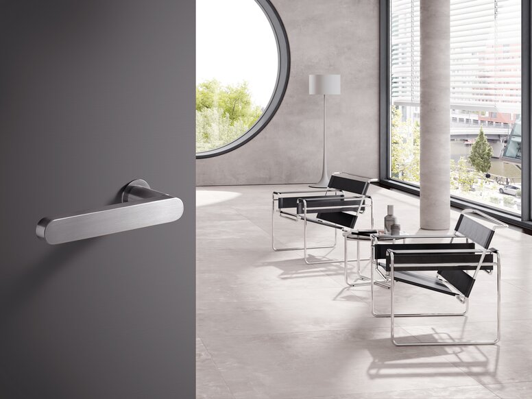 A stainless steel door handle on a grey door; behind it, a bright room with chairs