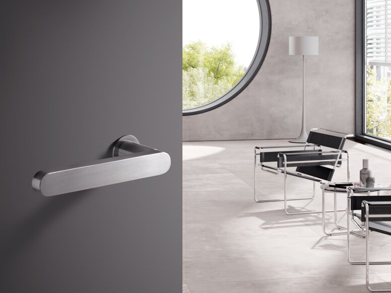 A stainless steel door handle on a grey door; behind it, a bright room with chairs