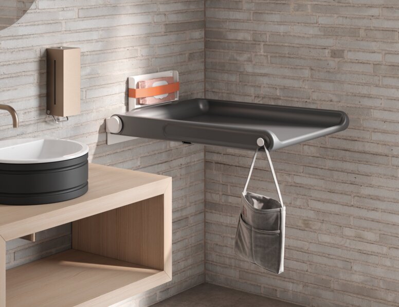 HEWI Range 480 Baby Changing Table A hinged baby changing table with a side pocket for storage and a hook, mounted on a light-coloured brick wall