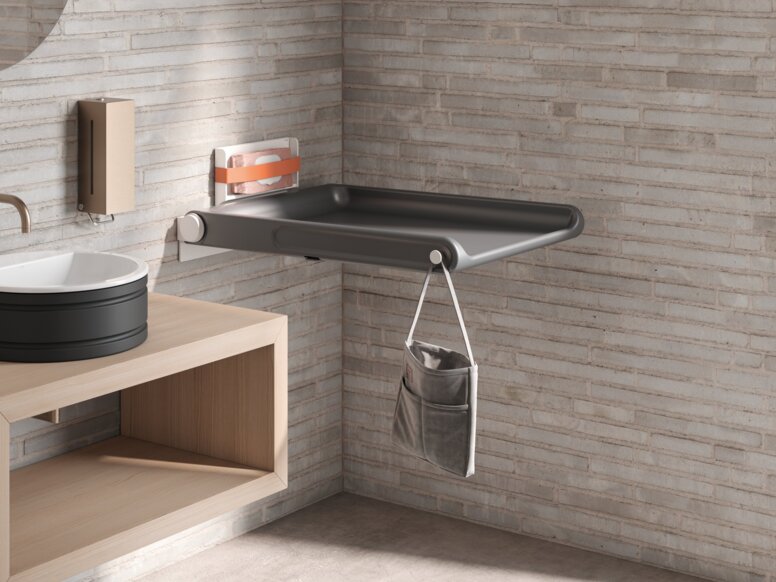 HEWI Range 480 Baby Changing Table A hinged baby changing table with a side pocket for storage and a hook, mounted on a light-coloured brick wall