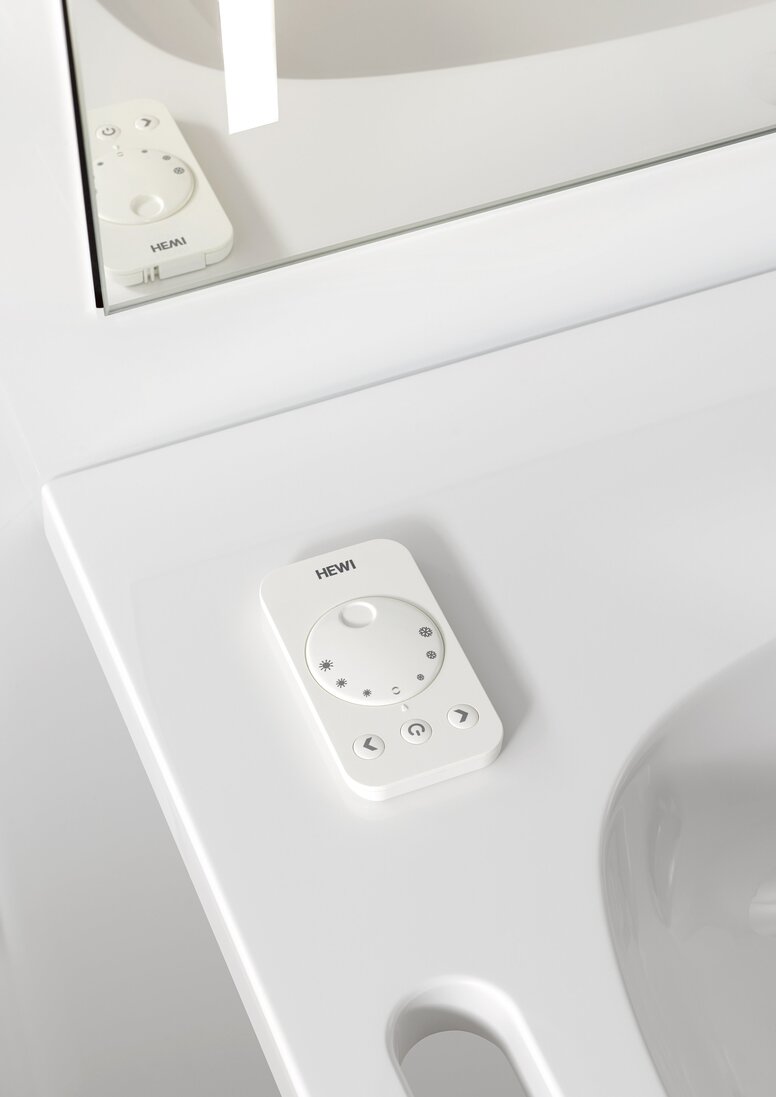 A white remote control with a dial, buttons and the HEWI logo, lying on the washbasin top