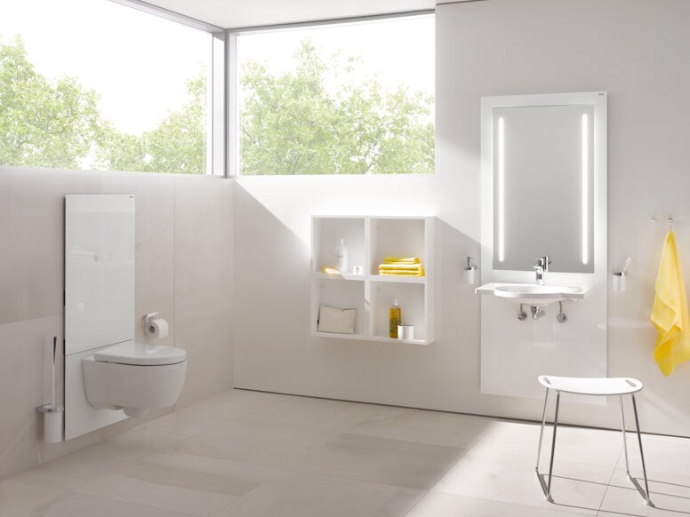 HEWI S 50 WC module A white WC module in a bright bathroom with an illuminated mirror above the washbasin, in front of which stands a stool with a white seat