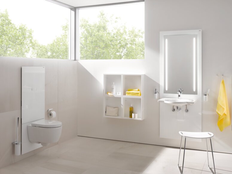 HEWI S 50 WC module A white WC module in a bright bathroom with an illuminated mirror above the washbasin, in front of which stands a stool with a white seat
