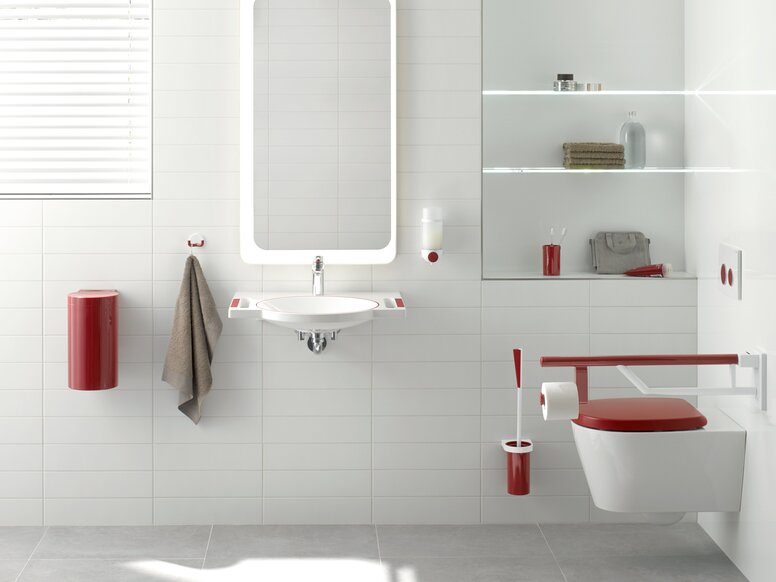 A red hinged support rail next to the WC with a red cover, alongside a white washbasin with red recessed handles, and an illuminated mirror above it