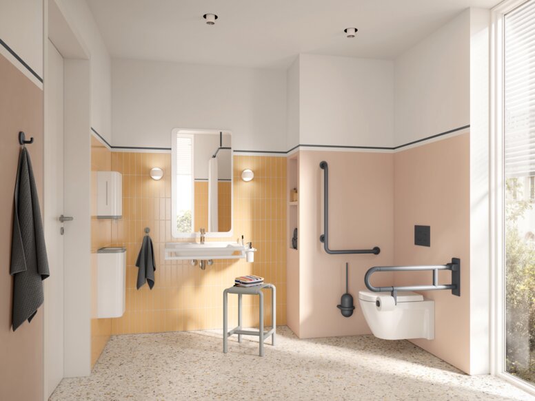HEWI Range 801 Interior Wall-hung WC with a hinged support rail on the side and an L-shaped support rail, alongside a washbasin with a mirror and a stool