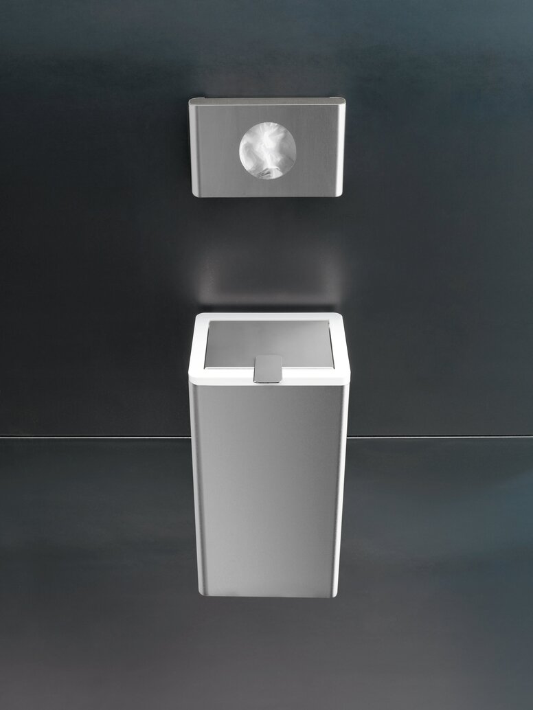 Rectangular stainless steel hygiene bag dispenser mounted above a matching waste bin on a dark grey wall