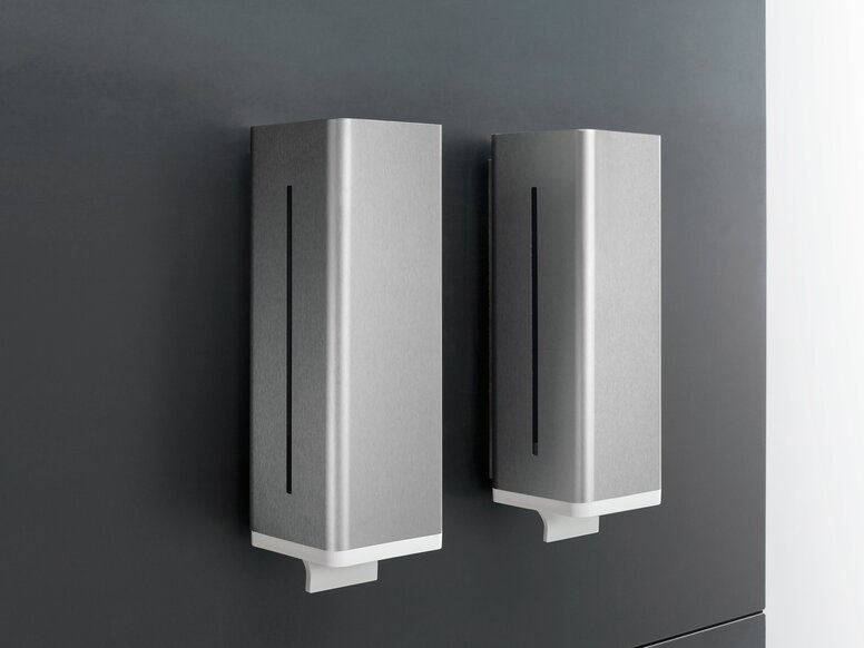 Two rectangular soap dispensers with silver-grey fronts, narrow vertical viewing windows and white dispensing outlets, mounted side by side on a dark grey wall