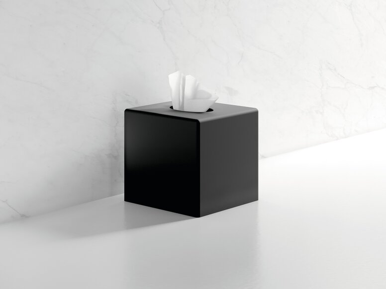 Black powder-coated cube-shaped tissue dispenser with an open cover and paper tissues, set against a white marble wall