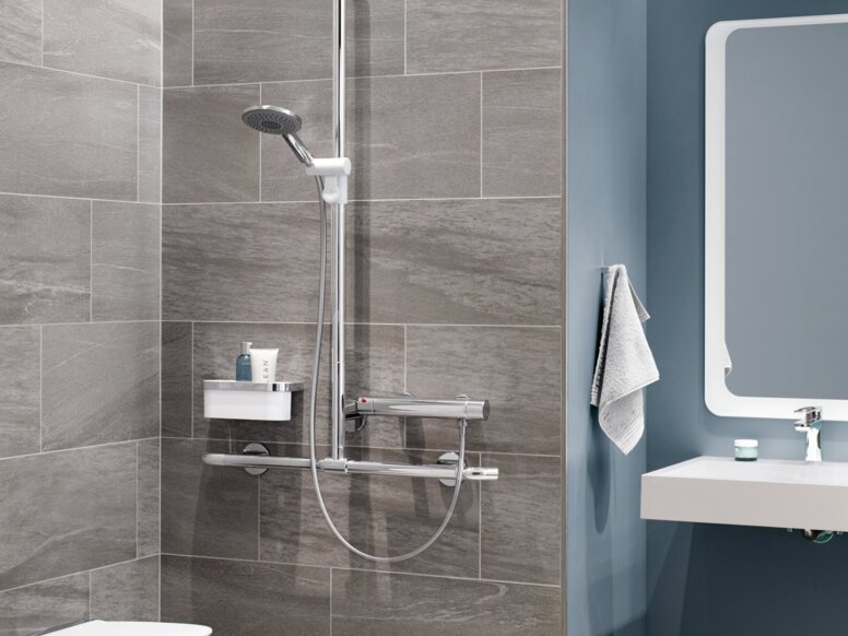 HEWI S950 Interior A hinged shower seat with a support rail, a thermostatic mixer and a shower head against a grey tiled wall, next to which is a washbasin area against a blue wall