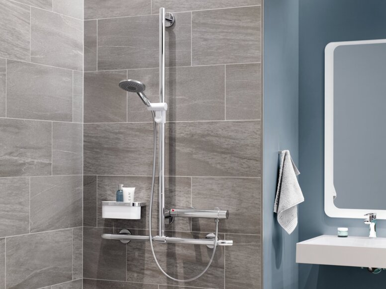 HEWI S950 Interior A hinged shower seat with a support rail, a thermostatic mixer and a shower head against a grey tiled wall, next to which is a washbasin area against a blue wall