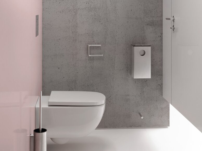 Narrow toilet area with a white wall-hung toilet against a grey concrete-effect wall, a white toilet roll holder and toilet brush set, and a pink side wall