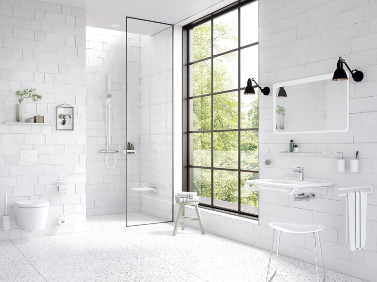 Bright bathroom with white wall tiles, a level-access shower behind a glass partition, a wall-hung toilet and a white washbasin beneath a mirror, complemented by white grab rails and a large black-framed grid window